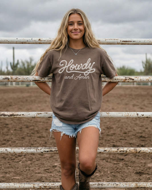 Howdy & Amen western cowgirl graphic tee in brown worn with denim shorts and cowboy boots at ranch fence