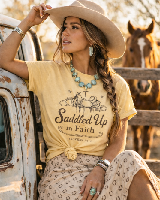 Cowgirl wearing Saddled Up in Faith western Christian graphic tee with horse in ranch background.