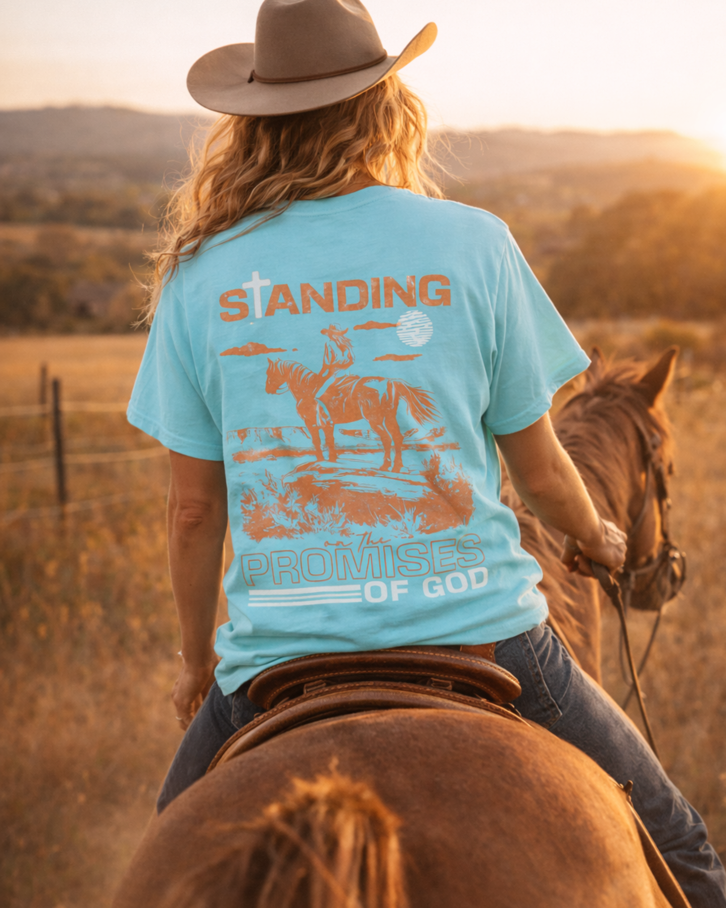Woman horseback riding wearing blue western faith graphic tee with back print, cowgirl outfit during golden hour in open pasture