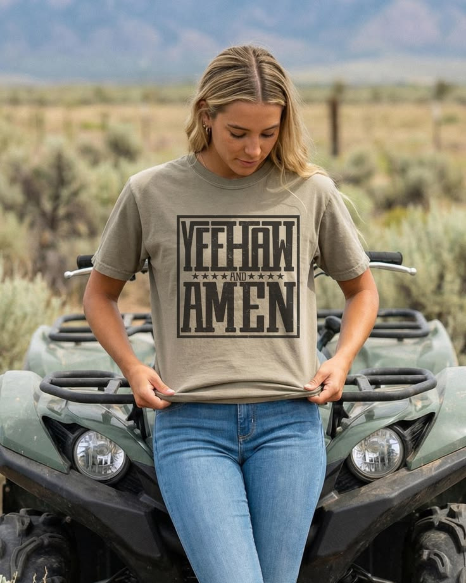 Woman wearing a Yeehaw and Amen Christian western graphic t-shirt sitting on an ATV in a desert landscape, styled with denim for a cowgirl outfit