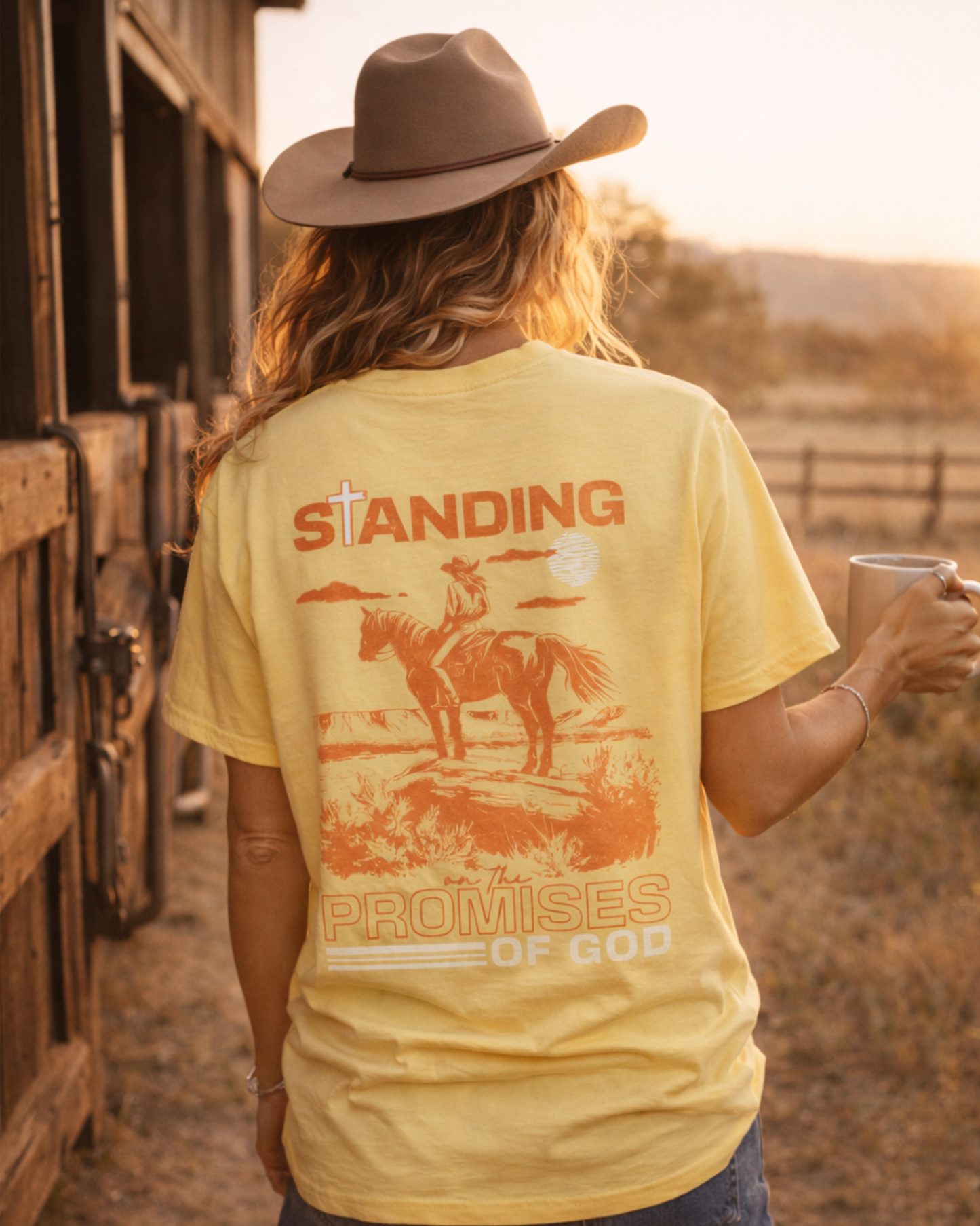 Woman wearing a yellow Christian cowgirl t-shirt with “Standing on the Promises of God” back graphic, holding coffee near a barn during golden hour